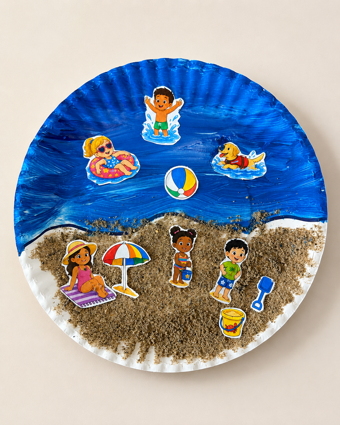 Beach small world play scene on a paper plate with printable people, beach toys, sand, and blue ocean paint